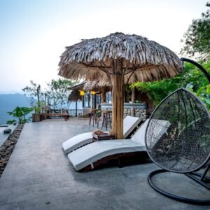 White loungers and thatched parasol with chair placed near swimming pool on terrace of tropical hotel with green exotic plants against mountain ridge on background