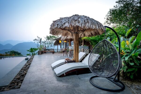 White loungers and thatched parasol with chair placed near swimming pool on terrace of tropical hotel with green exotic plants against mountain ridge on background